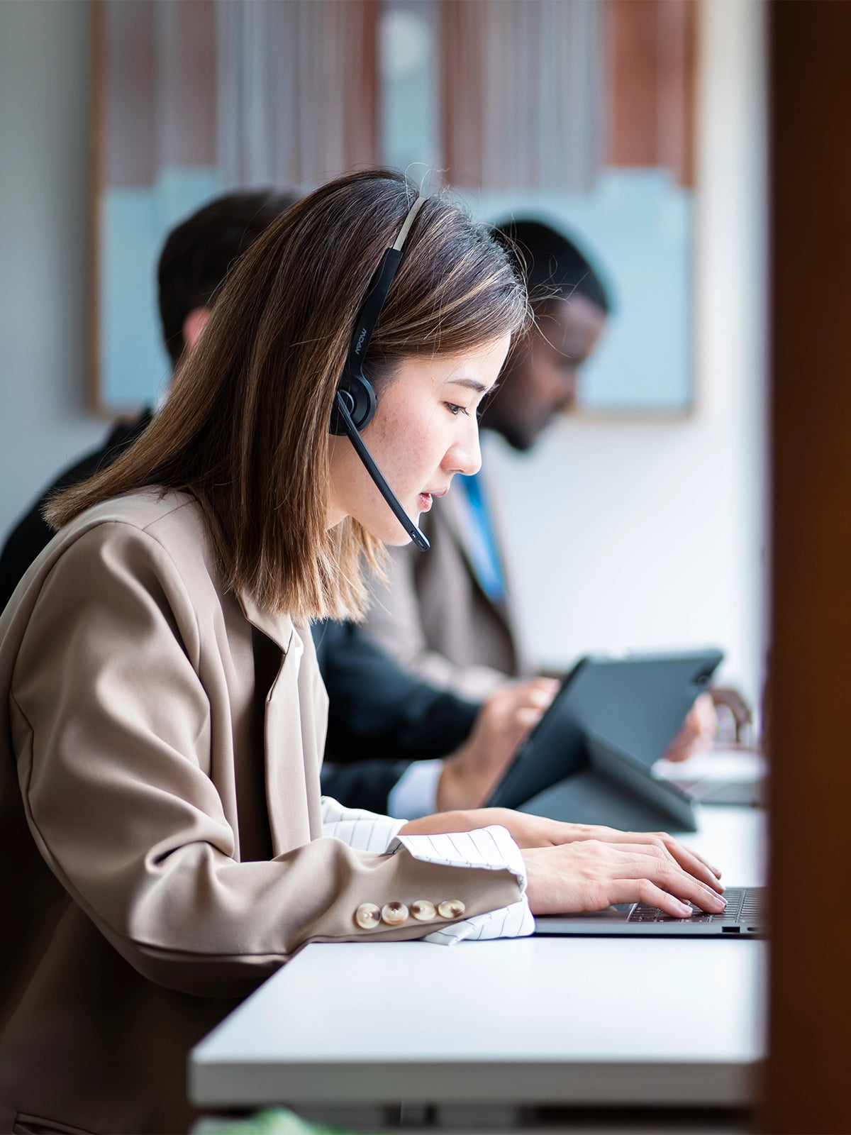 Person wearing a headset and using a laptop at a desk with colleagues in the background.