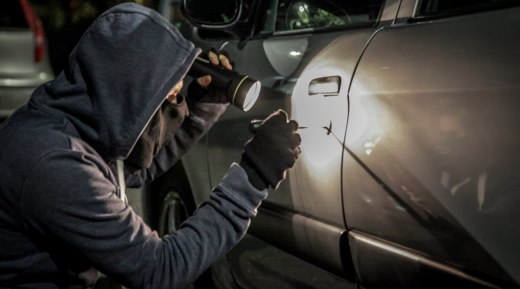 hooded man crouched next to car door at night with flashlight and screwdriver getting into car
